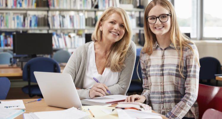 Kvinde og ung pige arbejder sammen ved et bord i et bibliotek med bøger og papirer foran sig.