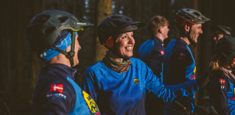 To kvinder med cykelhjem står i skov i aftenbelysning og smiler.