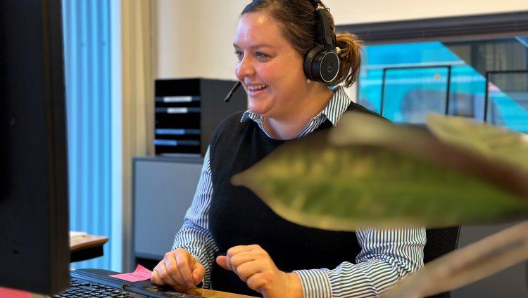 Kvinde med headset sidder i kontormiljø ved en computer og smiler. 